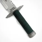 Rambo First Blood survival knife with a silver serrated blade, engraved signature, silver guard, and dark green wrapped handle on a white background with signature